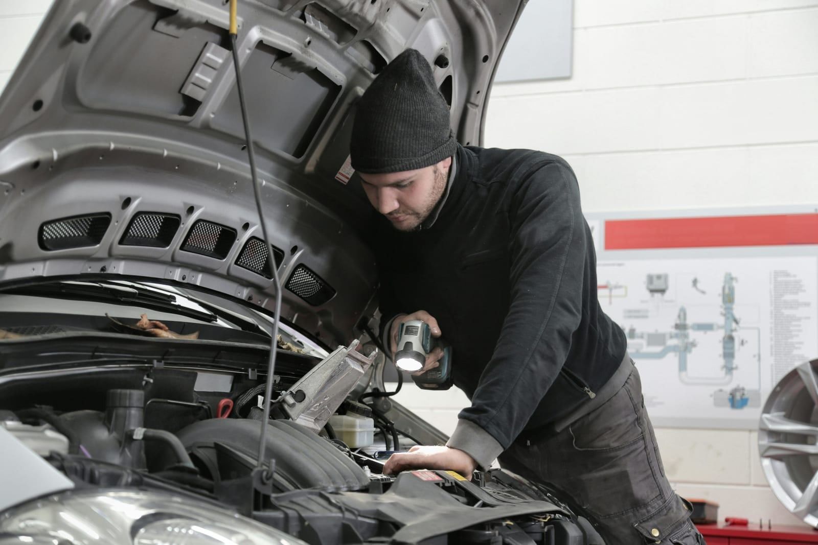 A mechanic in a beanie inspecting an engine bay with a work light.
