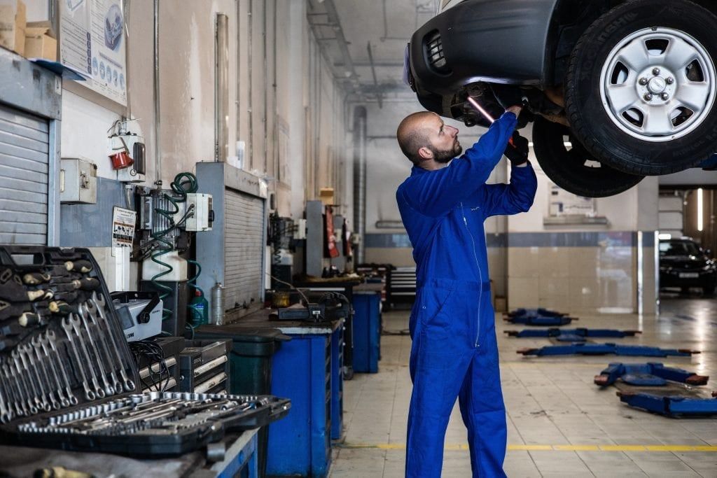 A mechanic in blue overalls working under a car raised on a lift in a busy repair shop.
