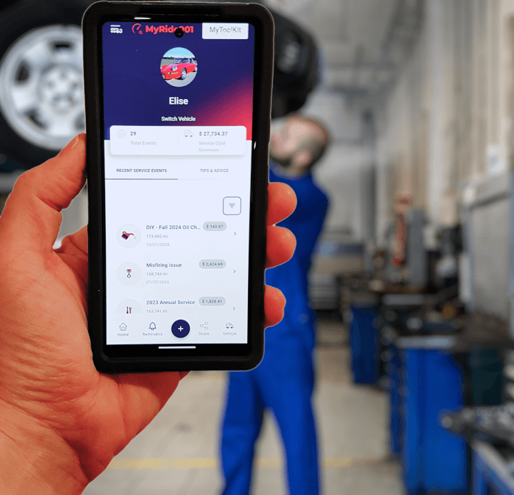 Hand holding a phone open on MyRide901, with a mechanic working under a car in the garage behind