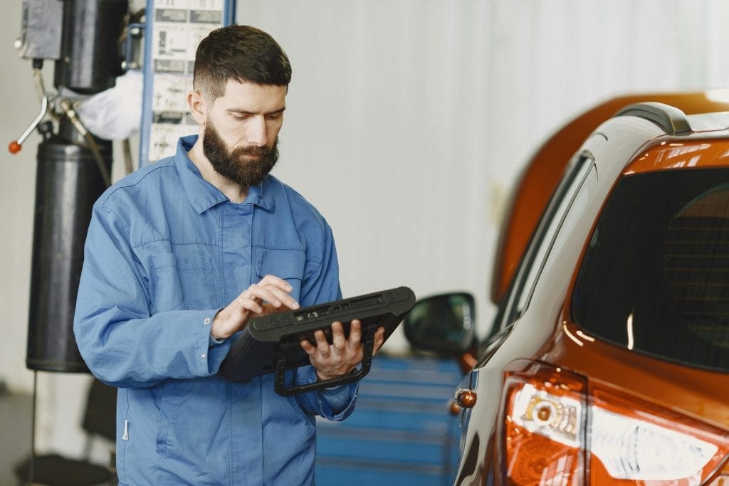 Mechanic reading a diagnostic tablet beside an orange car in a workshop