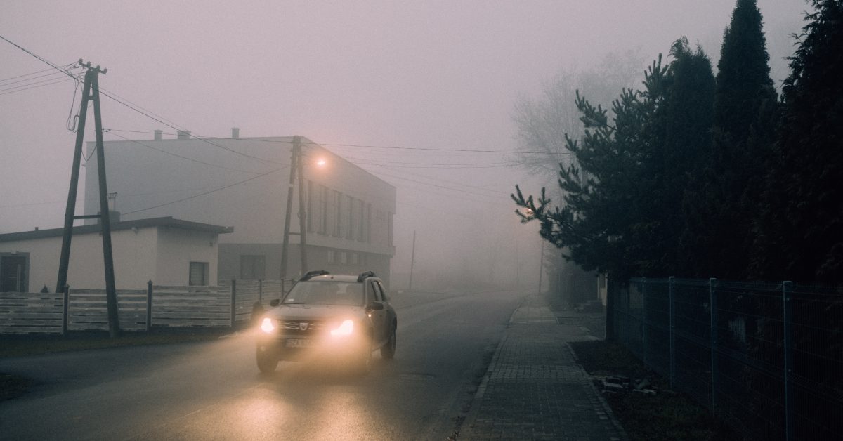 A car driving in on a roach with its headlights on in drizzly, low-visibility conditions.